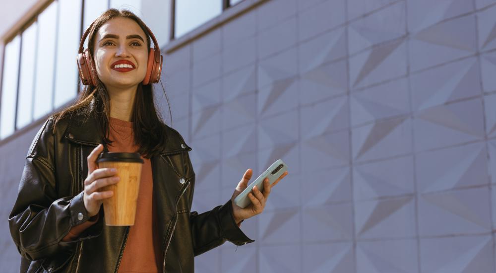 young smiling woman in headphones holding coffee and phone