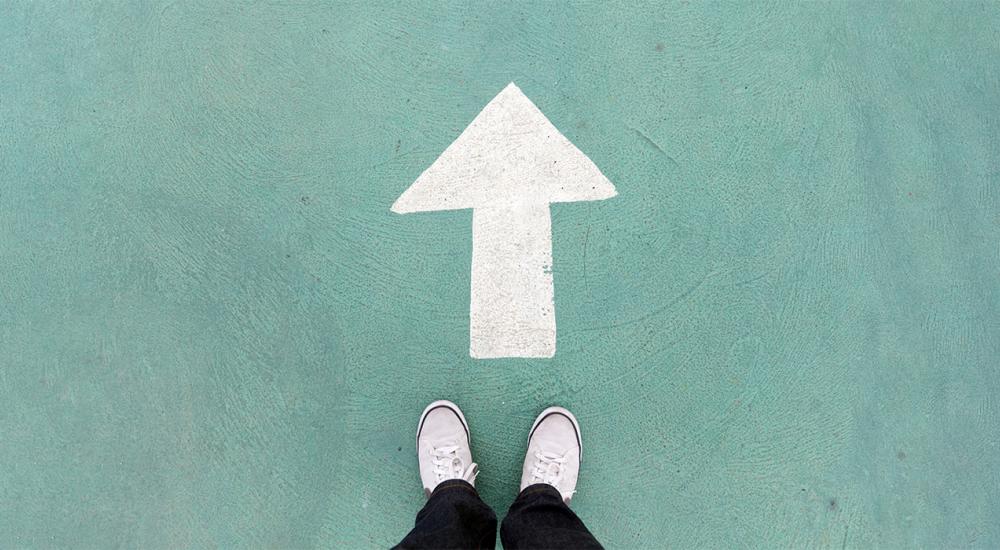 photograph of person's shoes as they satnd in front of upward arrow painted on turquoise colored background