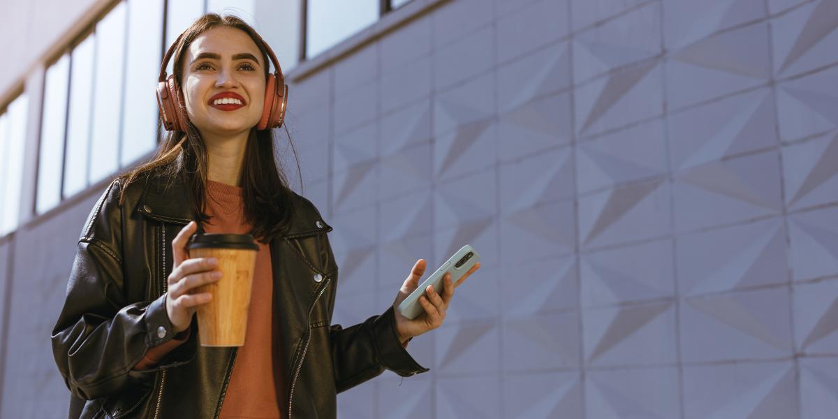 young smiling woman in headphones holding coffee and phone