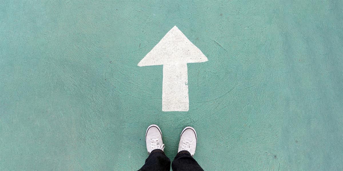 photograph of person's shoes as they satnd in front of upward arrow painted on turquoise colored background