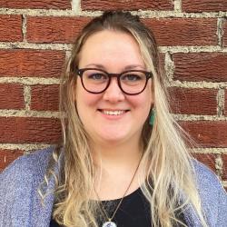 Headshot of Abby Conder. She is smiling at the camera, wearing glasses and leaning against a red brick wall.