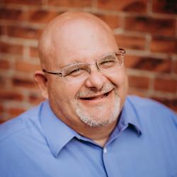 Peter Thomas Headshot. Peter smiles at the camera wearing glasses and a blue button up. Behind him is a brick wall out of focus. 