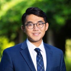 Man in a blue suit and tie, with black hair and glasses against a green background.