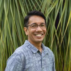 Man with short black hair and glasses wearing a patterned button-up shirt against palm trees in the background