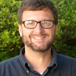 Man with short brown hair and beard, wearing glasses and dark polo against a green background