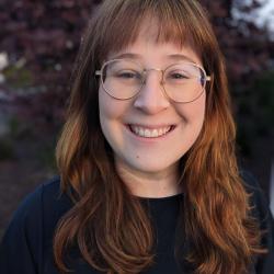 Woman with long brown hair wearing glasses and a dark top. Trees are in the background.