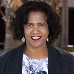 Woman with dark curly hair, wearing dangling earrings, purple and white top and a dark cardigan, against a dark background with shapes of trees.